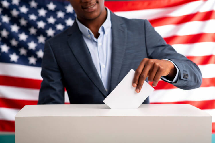 Em destaque, homem negro colocando voto na urna. Ao fundo, bandeira dos Estados Unidos da América.