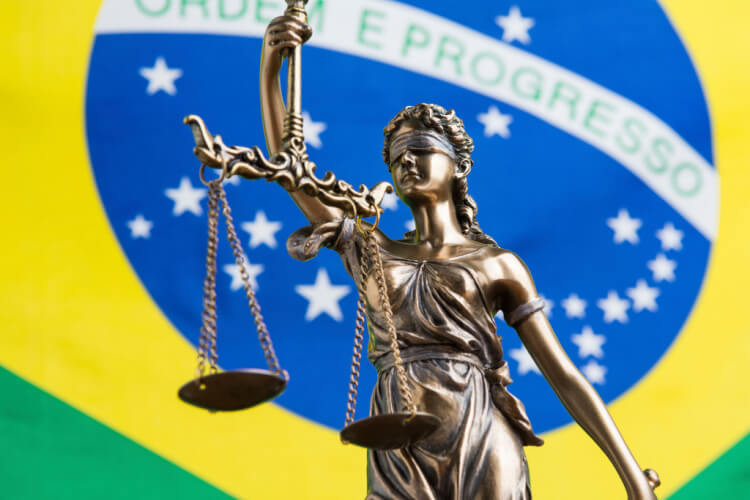 Em destaque, a estátua da Justiça, segurança a balança de dois pratos. Ao fundo, a bandeira do Brasil.