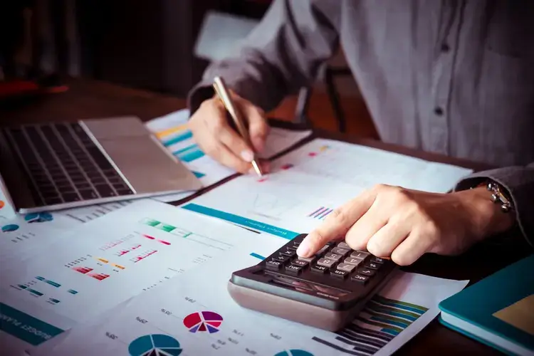 A imagem mostra uma pessoa fazendo a gestão financeira, com uma mesa cheia de documentos e uma calculadora sendo utilizada.